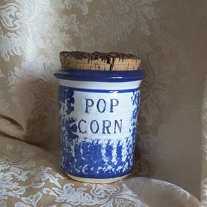 May include: Blue and white ceramic jar with a cork lid. The jar has a white glaze with blue speckles and the words "POP CORN" written in white.