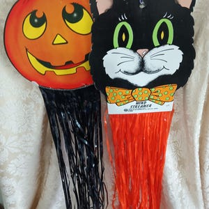 May include: Two Halloween decorations: a smiling orange pumpkin and a black cat with green eyes. Both have fringed tails and are hanging from a wire. The cat has a bow tie that says "Wind Streamer".