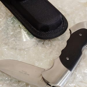 May include: A folding knife with a silver blade and a black handle, next to a black nylon sheath. The knife blade is partially open, displaying the text "Model 111D China". The knife has a stainless steel bolster.