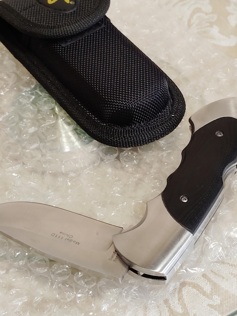 May include: A folding knife with a silver blade and a black handle, next to a black nylon sheath. The knife blade is partially open, displaying the text "Model 111D China". The knife has a stainless steel bolster.