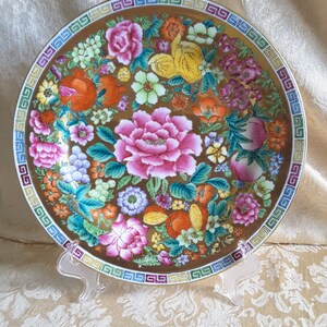 May include: A decorative plate with a gold background and a colorful floral design. The plate features pink peonies, orange fruits, and a border with a geometric pattern. The plate is displayed on a clear stand.