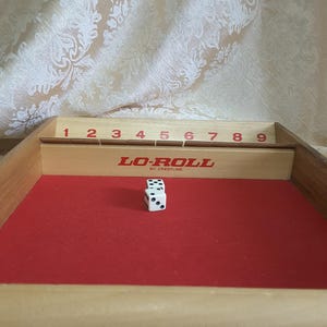 May include: Wooden Lo-Roll dice game with a red felt playing surface. The game features a wooden frame with numbers 1-9, and two white dice with black dots. The game is made by Crestline.