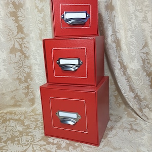 May include: Three stacked red square boxes with silver label holders and white stitching around the label area. The boxes are arranged in a pyramid shape, with the largest box at the bottom and the smallest at the top. The background is a patterned fabric.