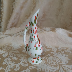 May include: A small white ceramic pitcher with a red and green floral pattern. The pitcher has a spout and a handle.