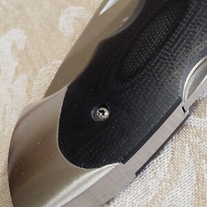 May include: Close-up of a partially open folding knife. The blade is silver, and the handle features a black textured grip with a silver bolster. A small screw secures the handle components. The knife rests on a beige patterned surface.