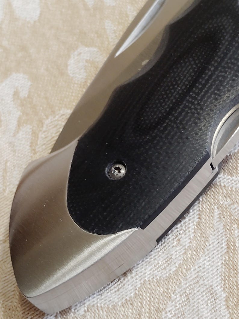 May include: Close-up of a partially open folding knife. The blade is silver, and the handle features a black textured grip with a silver bolster. A small screw secures the handle components. The knife rests on a beige patterned surface.