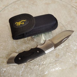 May include: A folding pocket knife with a stainless steel blade and a black and silver handle. The knife is next to a black nylon sheath with a yellow logo. The knife is approximately 20 cm long when open.