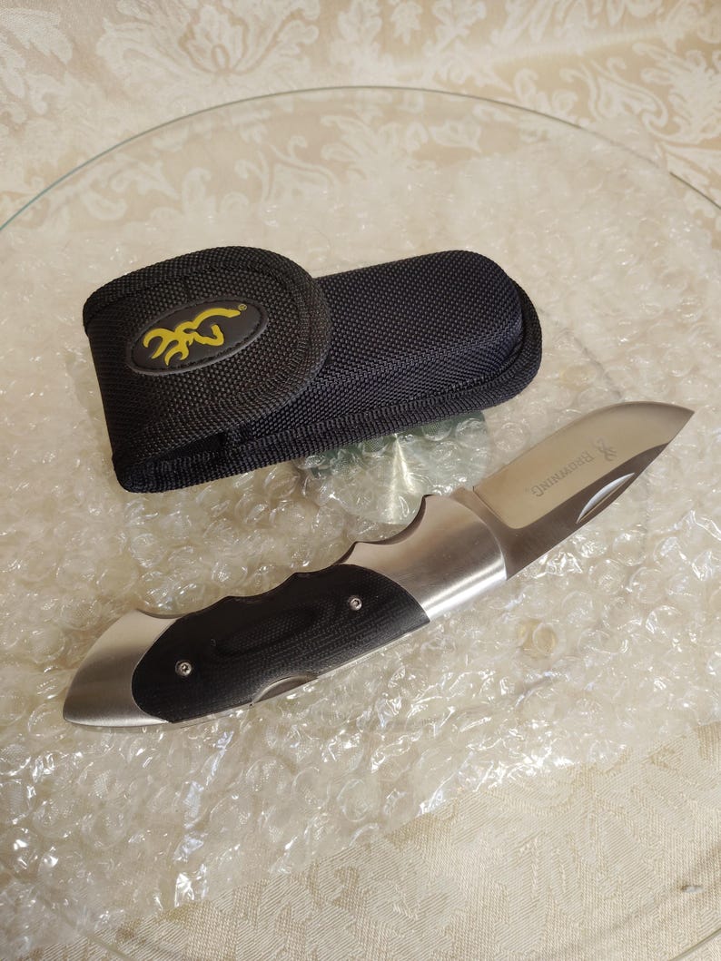 May include: A folding pocket knife with a stainless steel blade and a black and silver handle. The knife is next to a black nylon sheath with a yellow logo. The knife is approximately 20 cm long when open.