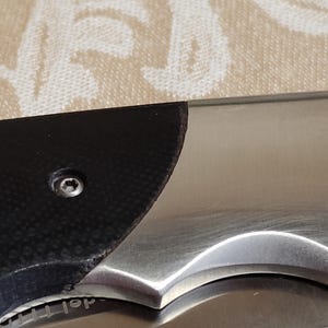 May include: Close-up of a partially visible folding knife. The blade is stainless steel, and the handle features a black textured grip. A small screw is visible on the handle. The knife is on a patterned surface.