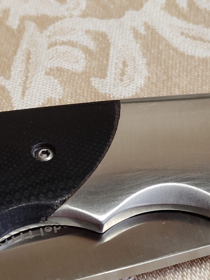 May include: Close-up of a partially visible folding knife. The blade is stainless steel, and the handle features a black textured grip. A small screw is visible on the handle. The knife is on a patterned surface.
