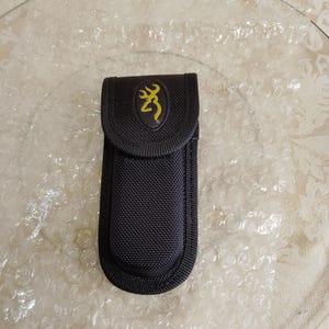 May include: Black nylon sheath with a yellow Browning logo on a black oval patch. The sheath is designed to hold a knife or tool. The sheath is on a clear glass plate.