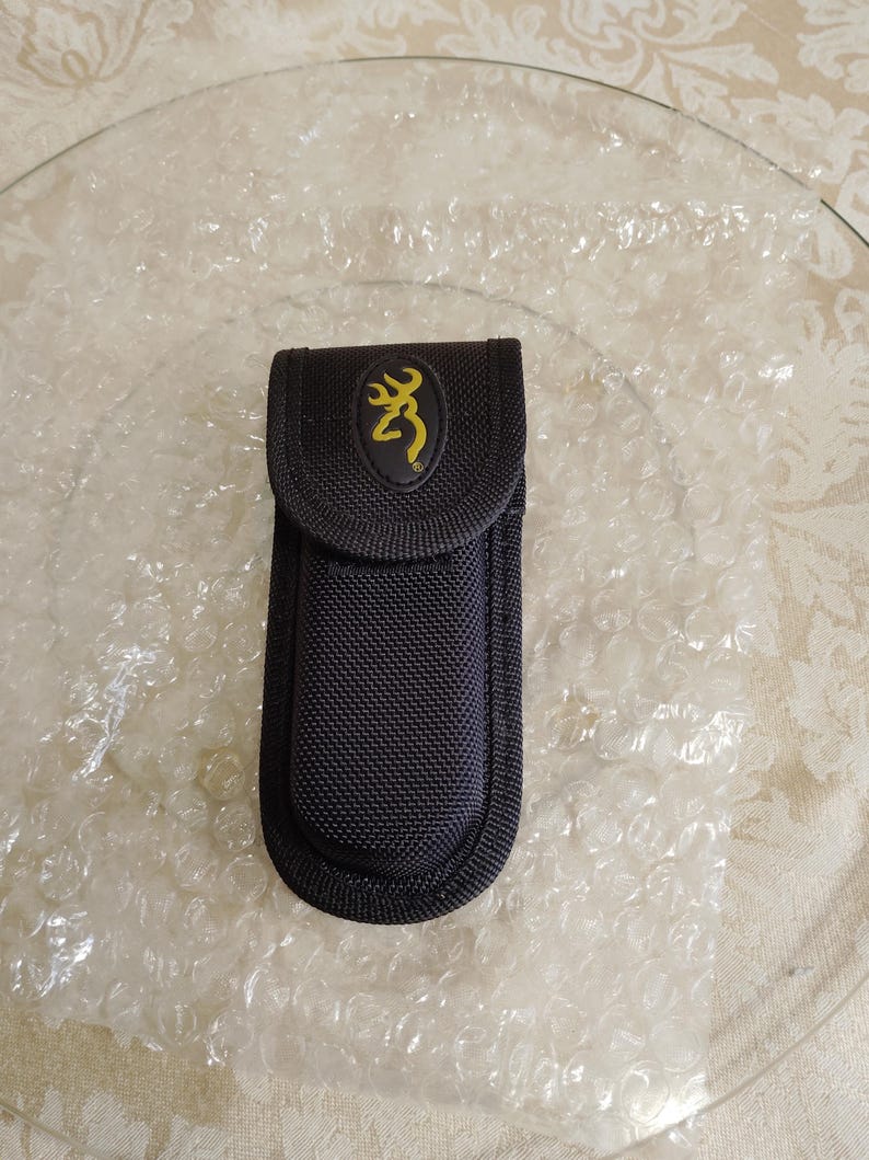 May include: Black nylon sheath with a yellow Browning logo on a black oval patch. The sheath is designed to hold a knife or tool. The sheath is on a clear glass plate.