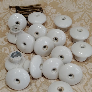 May include: A collection of white ceramic cabinet knobs with a smooth, rounded surface and a central hole for mounting. The set includes several metal screws with a dark, weathered finish, designed for attaching the knobs to furniture or doors.