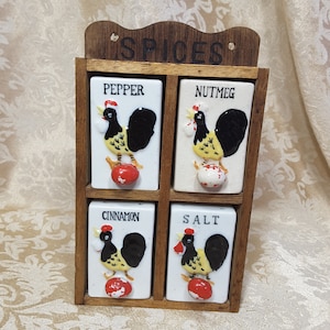 May include: A vintage spice rack with four ceramic spice containers. Each container is white with a black, yellow, and red rooster design. The containers are labeled Pepper, Nutmeg, Cinnamon, and Salt. The rack is made of wood and has the word "SPICES" at the top.