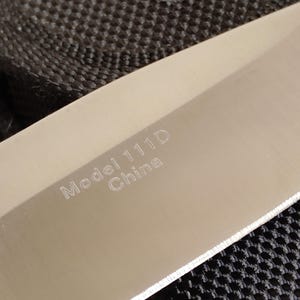 May include: Close-up of a stainless steel knife blade. The text "Model 111D China" is etched on the blade. The knife is set against a black textured background.