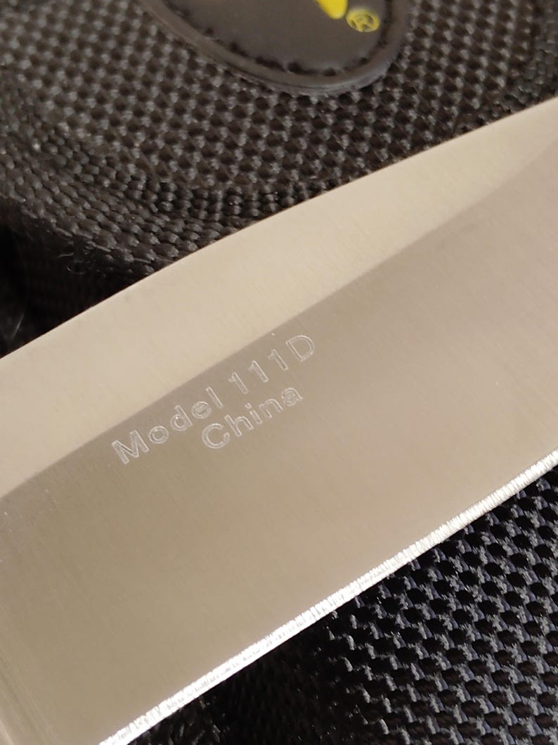 May include: Close-up of a stainless steel knife blade. The text "Model 111D China" is etched on the blade. The knife is set against a black textured background.