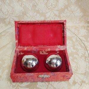 May include: A red, open box with a red interior holds two shiny, silver spheres. The box is decorated with a floral pattern and has a small clasp. The spheres reflect the surrounding environment.