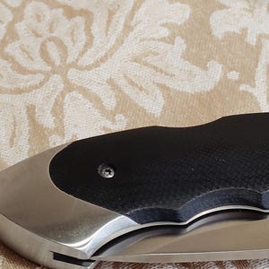 May include: A close-up of a folding knife. The knife has a stainless steel blade and handle. The handle has a black textured grip and a silver bolster. The knife is partially open, showing the sharp edge of the blade.