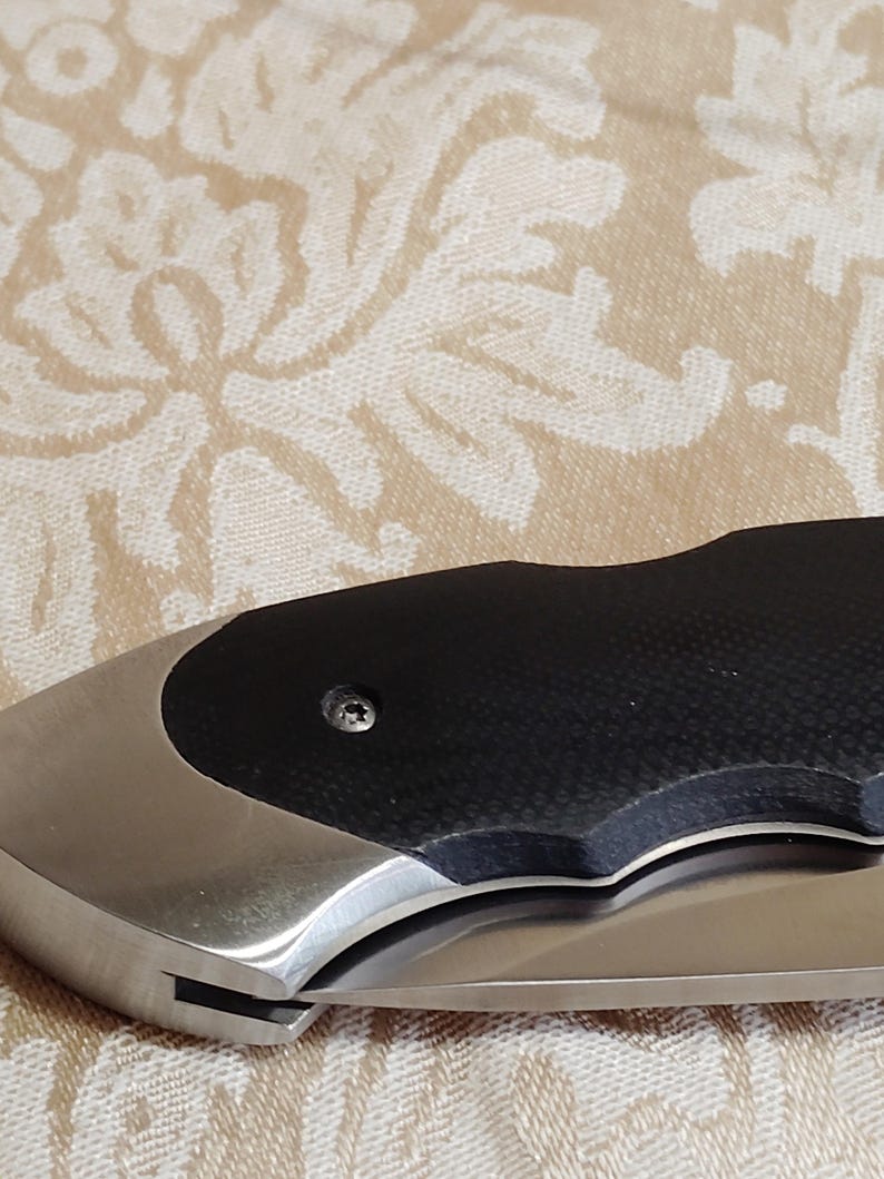 May include: A close-up of a folding knife. The knife has a stainless steel blade and handle. The handle has a black textured grip and a silver bolster. The knife is partially open, showing the sharp edge of the blade.