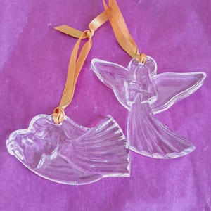 May include: Two clear glass angel ornaments with gold ribbon hangers. The angels have detailed wings and flowing gowns. The ornaments are displayed on a purple, textured background, suggesting a decorative or holiday theme.