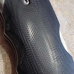 May include: Close-up of a knife handle with a textured, dark grey surface. The handle has a series of small, evenly spaced indentations. A silver metal component is visible near the top, with a small screw. The knife's blade is partially visible.