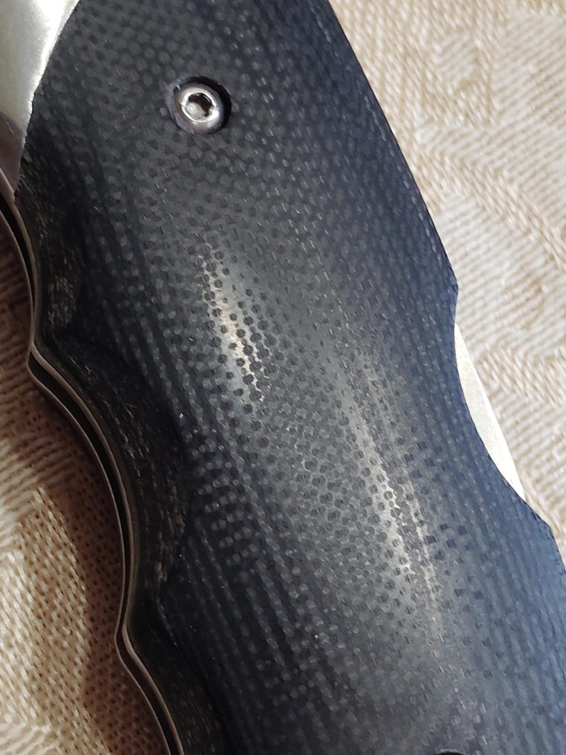 May include: Close-up of a knife handle with a textured, dark grey surface. The handle has a series of small, evenly spaced indentations. A silver metal component is visible near the top, with a small screw. The knife's blade is partially visible.