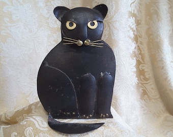 May include: A black metal cat sculpture with gold-colored eyes and whiskers. The cat is sitting upright with its tail curled. The sculpture has a vintage or antique appearance and is placed on a patterned fabric.