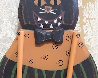 May include: A wooden Halloween cat decoration. The black cat has orange eyes, a wide grin with white teeth, and a black bow tie. The cat's body is orange with black swirls and green stripes. Orange wooden arms hang from the body.