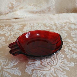 May include: A ruby red glass dish in the shape of a bird. The dish has a smooth, glossy finish and a dark rim. The bird's tail is fanned out, and the dish is resting on a patterned surface.