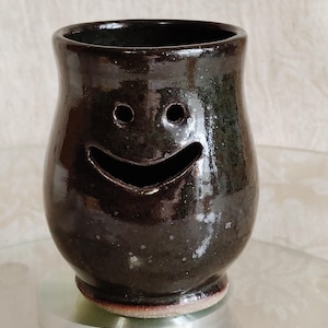 May include: A handmade, dark brown ceramic vase with a smiling face design. The vase has two eye holes and a wide, curved mouth. The glossy finish reflects light, highlighting the unique shape and texture.
