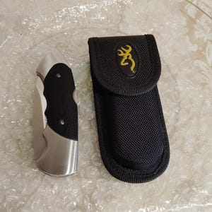 May include: A stainless steel folding knife with a black handle and a black nylon sheath. The sheath features a yellow deer logo. The knife and sheath are on a clear, textured surface.