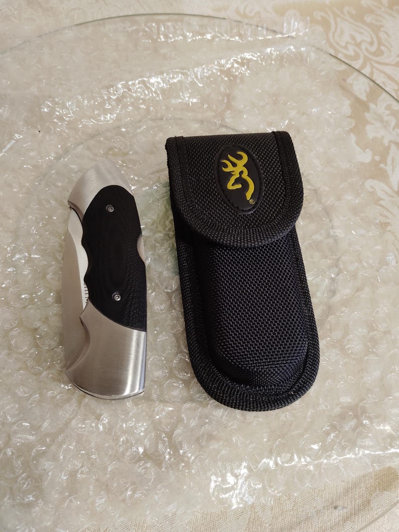 May include: A stainless steel folding knife with a black handle and a black nylon sheath. The sheath features a yellow deer logo. The knife and sheath are on a clear, textured surface.