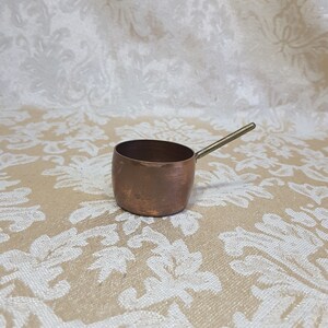 May include: A small copper saucepan with a brass handle. The pan is round with a short, straight side and a long handle. The copper has a warm, reddish-brown color.