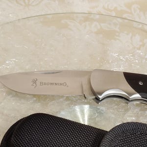 May include: A stainless steel folding knife with a dark wood handle. The blade is engraved with the Browning logo. A black nylon carrying case is included.