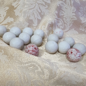 May include: A collection of small, round, white objects, some with red speckled patterns, are arranged on a textured, cream-colored fabric. The objects appear to be decorative or possibly edible, creating a visually interesting display.
