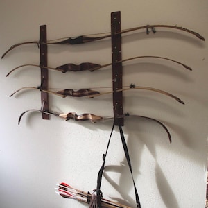 May include: Five wooden bows are mounted on a wall rack with brown leather straps. The bows are arranged horizontally, with the top bow being the shortest and the bottom bow being the longest. A black strap hangs from the bottom bow, and a quiver of arrows is on the floor below the rack.