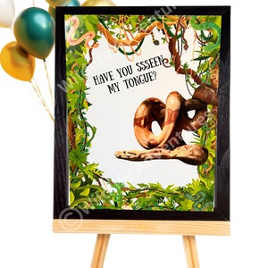 May include: A framed print on a wooden easel with a snake illustration. The artwork includes the text "HAVE YOU SSSSEEN MY TONGUE?" surrounded by jungle foliage and vines. Balloons in gold, green, and white are in the background.