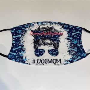 May include: A white face mask with a blue and white design featuring a woman with a messy bun and sunglasses. The text "#DOGMOM" is printed on the mask.