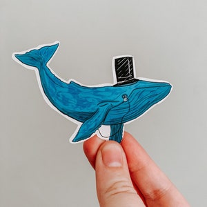 May include: A blue whale sticker wearing a black top hat. The whale has a detailed design with shading and highlights. The sticker is held between fingers against a neutral background. The whale has a small earbud.