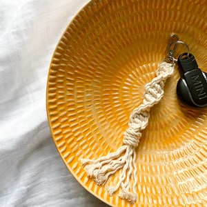 May include: A white macrame keychain with a black car key fob that says "MINI" on it. The keychain is sitting on a yellow ceramic plate with a textured pattern.