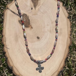 Puede incluir: Un collar de cuentas con un colgante de cruz plateada. El collar presenta cuentas ovaladas en tonos beige e iridiscentes, intercaladas con cuentas más pequeñas y coloridas. El colgante de cruz tiene una superficie texturizada.