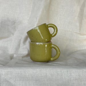 Puede incluir: Dos tazas pequeñas de cerámica verde oliva apiladas una encima de la otra. Cada taza tiene un asa redondeada y una superficie ligeramente texturizada. Las tazas están sobre un fondo blanco texturizado.