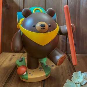 May include: A brown bear figurine with a yellow scarf and a blue backpack, holding a red stick. The bear is standing on a green and brown base with a mushroom.