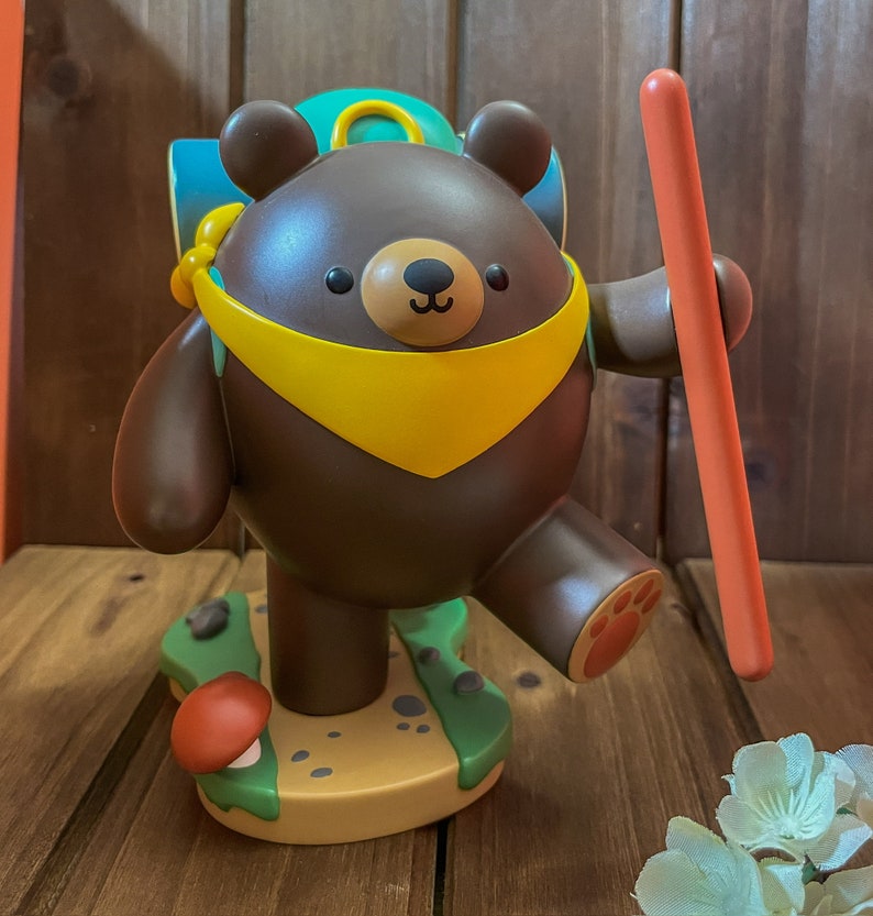 May include: A brown bear figurine with a yellow scarf and a blue backpack, holding a red stick. The bear is standing on a green and brown base with a mushroom.