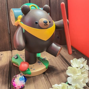 May include: A brown bear figurine with a yellow scarf and a green backpack, holding a red stick. The bear is standing on a green and brown base with a red mushroom. A multi-coloured dice is in the foreground.
