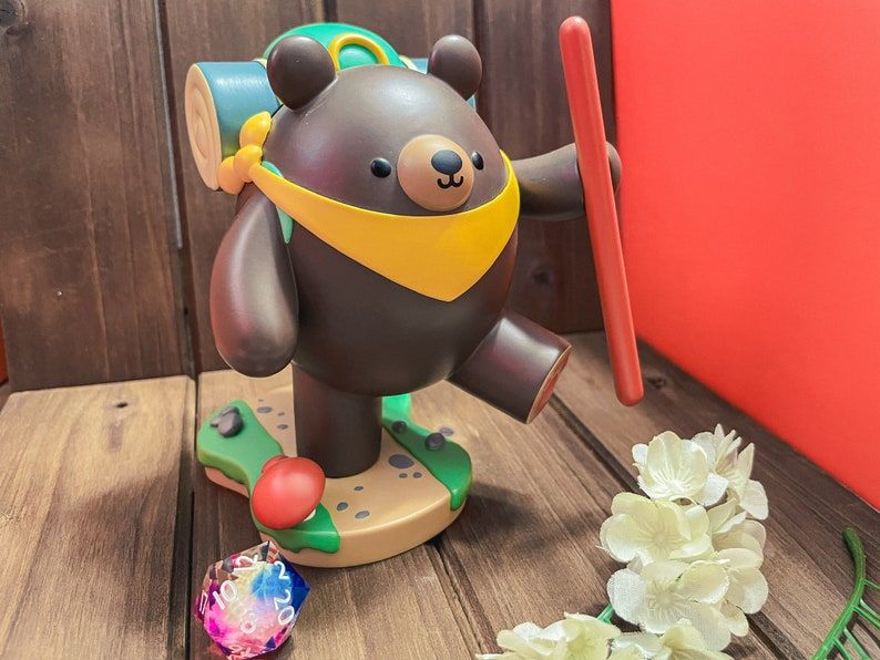 May include: A brown bear figurine with a yellow scarf and a green backpack, holding a red stick. The bear is standing on a green and brown base with a red mushroom. A multi-coloured dice is in the foreground.