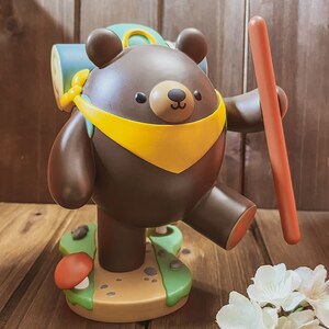 May include: A brown bear figurine with a yellow scarf and a blue backpack, holding a red stick. The bear is standing on a green and brown base with a mushroom.