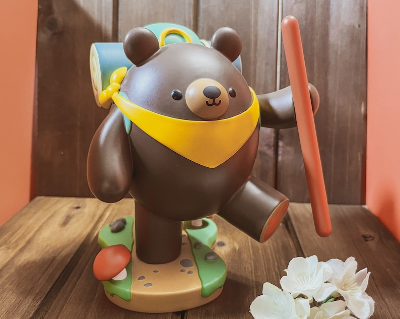 May include: A brown bear figurine with a yellow scarf and a blue backpack, holding a red stick. The bear is standing on a green and brown base with a mushroom.