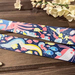 May include: Navy blue ribbon featuring a colorful underwater scene with various sea creatures. The design includes an octopus, fish, jellyfish, and other marine life in shades of pink, yellow, and blue. The ribbon is likely used for crafting or gift wrapping.