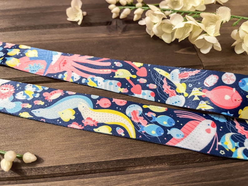 May include: Navy blue ribbon featuring a colorful underwater scene with various sea creatures. The design includes an octopus, fish, jellyfish, and other marine life in shades of pink, yellow, and blue. The ribbon is likely used for crafting or gift wrapping.